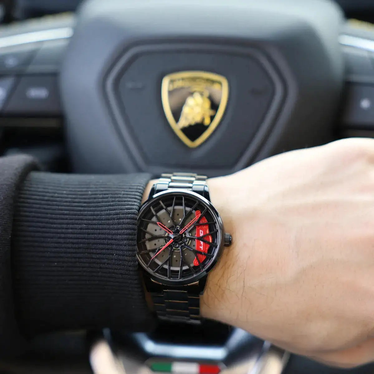 Black motorsports watch with red accents on wrist against a racing backdrop.